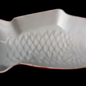 Op de afbeelding: Een witte keramische serveerschaal in de vorm van een vis, voorzien van een gegoten schubbenpatroon aan de binnenzijde en een dunne, roodbruin geschilderde rand, ontworpen met een brede staart en een afgerond kopgedeelte.