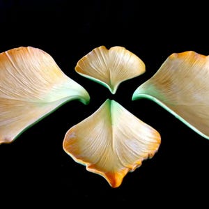 Op de afbeelding: Vier gestileerde, bladvormige decoratieve objecten in tinten geel, oranje en groen zijn geplaatst tegen een zwarte achtergrond. De objecten hebben een textuur, met zichtbare aderingdetails. Het algehele ontwerp suggereert een botanisch thema.