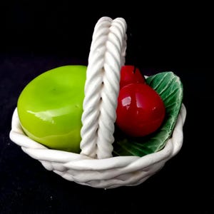 Puede incluir: Una cesta de cerámica blanca con asa trenzada que contiene una manzana verde lima brillante, una cereza roja reluciente y una hoja verde texturizada. La cesta presenta un borde y una base de estilo tejido como recipiente decorativo.