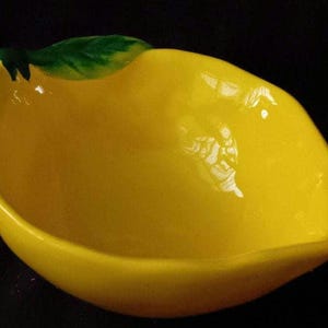 Könnte beinhalten: Eine zitronenförmige Keramikschale mit glänzend gelbem Finish. Die Schale hat einen grünen Blattakzent auf der Oberseite und ist mit einer flachen Schalenform gestaltet. Die leuchtend gelbe Farbe und die einzigartige Form machen es zu einem dekorativen Stück.