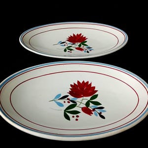 May include: Two white oval serving platters featuring a central floral motif with a large red flower, green leaves, and small blue accents. The rim of each platter is decorated with a thin red line and a wider light blue border line.
