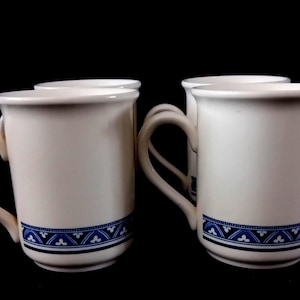 May include: Four white ceramic mugs with a blue and black geometric pattern around the base. Each mug has a curved handle and a slightly flared rim. The mugs are set against a black background.