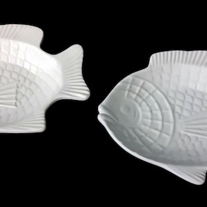 Pode incluir: Dois pratos de cerâmica branca em forma de peixe, apresentando padrões de escamas em relevo no corpo, um design circular na cabeça e barbatanas detalhadas. Os pratos têm um acabamento liso e brilhante sobre um fundo preto sólido.