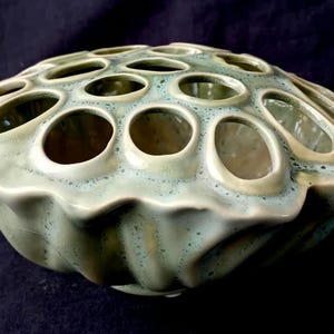 Op de afbeelding: Een keramische bloemenkikker met een zeegroene glazuur. De kikker heeft een afgeronde, gerimpelde basis en een bovenoppervlak met talrijke cirkelvormige openingen. De openingen zijn ontworpen om bloemstelen vast te houden, waardoor het een decoratief en functioneel stuk is.