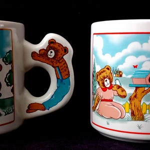 Può includere: Due tazze in ceramica bianca con illustrazioni colorate di orsacchiotti marroni in ambienti esterni. Ogni tazza ha un manico unico a forma di orso. I disegni raffigurano orsi con pompe dell'acqua, casette per uccelli e recinzioni.