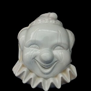 Peut inclure: Une tête de clown en céramique blanche avec une expression souriante, un col à volants et un petit chapeau. Le clown a des détails peints, dont un nez rouge et des lignes peintes sur les joues. Le fond est noir.