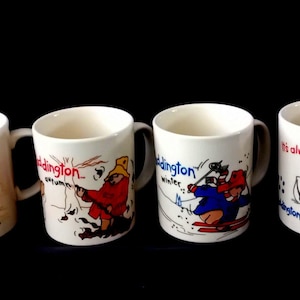 Op de afbeelding: Zes witte keramische mokken met Paddington Bear-illustraties. Elke mok toont een andere scène van Paddington Bear in verschillende activiteiten, met de naam "Paddington" in rode letters. De mokken hebben een standaard handvat en een glanzende afwerking.