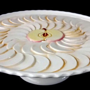 Op de afbeelding: Witte keramische serveerschaal met een decoratief appelontwerp. De schaal heeft een geschulpte rand en een centraal appelontwerp gemaakt van witte en roze keramische stukken. De appelschijfjes zijn in een spiraalvorm gerangschikt.