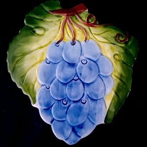 Op de afbeelding: Een decoratieve keramische plaat in de vorm van een druivenblad, met een tros blauwe druiven. De druiven zijn vastgebonden met een rood lint. Het blad is groen met gele accenten, tegen een zwarte achtergrond.