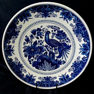 May include: A white ceramic plate with a blue floral and bird design. The plate features a central image of a bird surrounded by flowers and foliage. The rim of the plate is decorated with a repeating floral pattern. The plate is displayed on a black stand.