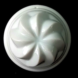 Op de afbeelding: Een ronde witte keramische vorm met een verhoogd, wervelend, windmolenachtig patroon dat vanuit het midden uitstraalt. Het object heeft een gladde, glanzende afwerking en een duidelijke, afgeronde rand rondom het ontwerp.
