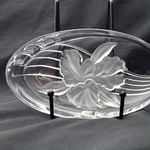 Peut inclure: Plat ovale en verre transparent avec un motif de fleur givrée. Le plat est soutenu par un support en métal noir. La fleur est au centre du plat et présente des pétales et des feuilles détaillés.