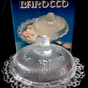 Op de afbeelding: Een heldere glazen snoepschaal met een deksel, met een getextureerd oppervlak en bloemaccenten. De schaal heeft een kralenrand en wordt gepresenteerd voor een blauwe doos met het woord "BAROCCO".