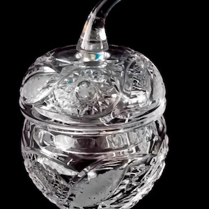 Op de afbeelding: Een heldere, geslepen glazen pot met een deksel en een gebogen handvat. De pot heeft een ronde vorm met ingewikkelde patronen en ontwerpen. Het deksel past perfect op de bovenkant en het handvat buigt omhoog. De pot staat tegen een zwarte achtergrond.