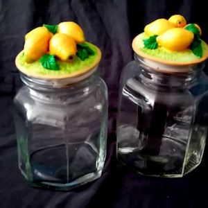 Op de afbeelding: Twee doorzichtige glazen potten met zeshoekige lichamen en deksels. Elk deksel heeft een getextureerde groene basis bekroond met gebeeldhouwde gele citroenen en groene bladeren. De potten zijn leeg en hebben een transparante afwerking.