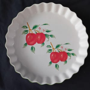 Op de afbeelding: Witte keramische taartschotel met een geschulpte rand en een groene rand. De schotel heeft een ontwerp van rode appels met groene bladeren en witte bloemen. Een decoratieve serveerschaal.