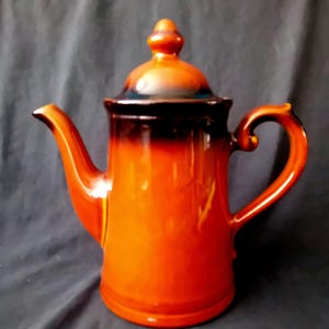 Op de afbeelding: Een keramische koffiepot met een glanzende afwerking, voorzien van een feloranje basis die naar boven toe overgaat in een donkerbruin glazuur. De pot heeft een bijpassend deksel met een knop, een gebogen tuit en een stevig zijhandvat.