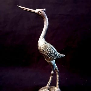 Op de afbeelding: Zilverkleurig reigerbeeldje met een lange, gebogen nek en gedetailleerde bevedering. De vogel staat op een ronde basis, zijn poten en snavel zijn slank en puntig. Het beeldje is een decoratief item.