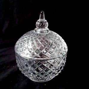 Op de afbeelding: Een heldere, ronde glazen bonbonnière met een ruitpatroon. De schaal heeft een deksel met een decoratieve bekroning. Het glas is transparant, waardoor licht erdoorheen kan schijnen, en staat tegen een zwarte achtergrond.