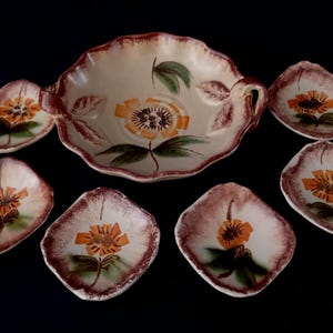 Op de afbeelding: Een vintage keramische serveerset met een grote kom en zes kleinere schaaltjes. Elk stuk is beschilderd met een bloemmotief in oranje, bruin en groen op een crèmekleurige achtergrond. De set heeft een geschulpte rand.