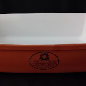 Op de afbeelding: Rechthoekige ovenschaal met een witte binnenkant en een terracotta-kleurige buitenkant. De schaal heeft een licht uitlopende rand en kleine handvatten aan elk uiteinde. Een decoratief embleem staat op de voorkant.