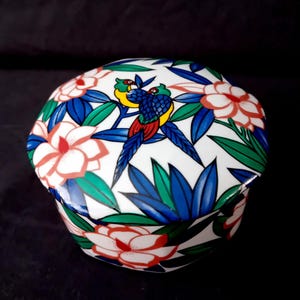 Op de afbeelding: Witte keramische sieradendoos met een bloemen- en vogelontwerp. Het deksel toont twee papegaaien met blauwe, gele en rode veren, omringd door blauwe bladeren en roze en witte bloemen. De doos heeft een geschulpte rand.