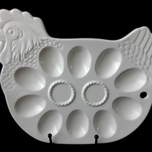 May include: A white ceramic egg tray shaped like a chicken, featuring twelve oval indentations for holding eggs. The design includes a detailed head and tail, with two central circular rings and a small hole on the right side for hanging.