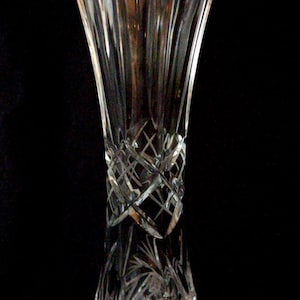 Op de afbeelding: Een heldere kristallen vaas met een uitlopende bovenkant en een diamantgeslepen patroon in het midden. De vaas heeft verticale lijnen en ingewikkelde stervormige ontwerpen aan de basis, tegen een zwarte achtergrond.