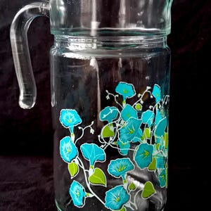 Op de afbeelding: Transparante glazen kan met een gebogen handvat en een schenktuit. De kan is versierd met een bloemenontwerp met turquoise bloemen en groene bladeren. De kan is geschikt voor het serveren van dranken.