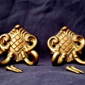 Peut inclure: Quatre patères en métal doré, chacune présentant un motif de grille carrée centrale entouré de volutes ornementales. Chaque patère possède deux bras incurvés et deux trous de fixation. Quatre petites vis en métal doré sont incluses.