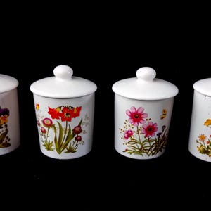 Op de afbeelding: Vier witte keramische potten met deksels, elk versierd met een ander bloemenontwerp. De bloemen omvatten viooltjes, klaprozen, roze madeliefjes en oranje bloemen. De potten zijn cilindrisch met een kleine knop bovenop.