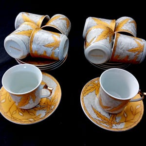 Op de afbeelding: Een set porseleinen kopjes en schotels. De kopjes en schotels hebben een gouden bladeren ontwerp op een witte en grijze achtergrond. De schotels zijn geel met een gouden bladeren rand. De kopjes hebben gouden handvatten.