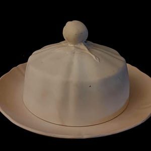 Op de afbeelding: Een witte keramische botervloot met een gewelfde deksel en een ronde knop. De schaal heeft een geschulpte rand en staat op een bijpassend bord. Een klassiek keukenaccessoire.