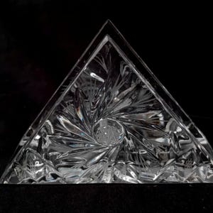 Peut inclure: Un porte-serviettes en cristal clair de forme triangulaire avec des motifs gravés complexes. Le design présente une spirale centrale et des lignes rayonnantes, créant un effet d'éclat. Les bords sont nets et la forme est géométrique.