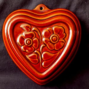 Op de afbeelding: Een hartvormige keramische wandplaat met een glanzende, diep oranjebruine glazuurlaag. Het midden is voorzien van een verhoogd, reliëf ontwerp van twee gestileerde bloemen met bladeren en stengels. Een klein gat bevindt zich bovenaan in het midden.