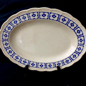 Op de afbeelding: Ovale serveerschaal met een crèmekleurige basis en een blauwe bloemenrand. De rand heeft een herhalend patroon van blauwe bloemachtige vormen. De schaal heeft een geschulpte rand en wordt op een zwarte standaard gepresenteerd.