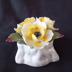 May include: A ceramic floral arrangement featuring yellow and white flowers with a dark center, set in a white vase. The leaves are green with red tips. The arrangement is a decorative piece.