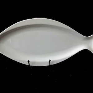 Könnte beinhalten: Eine weiße Servierplatte aus Keramik in Fischform. Die Platte hat eine glatte, glänzende Oberfläche mit einem spitz zulaufenden Kopf und einer gegabelten Schwanzflosse. Sie steht auf einem kleinen schwarzen Metallständer und zeigt ihre Form.