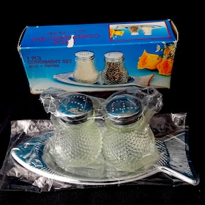 Op de afbeelding: Een tweedelige kruidenset in een blauwe doos. De set bevat twee glazen zout- en peperstrooiers met blauwe deksels, op een visvormige schaal. De doos leest "2 PCS CONDIMENT SET SALT + PEPPER".