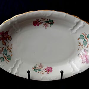 Op de afbeelding: Een ovale witte porseleinen serveerschaal met een geschulpte rand en een goudkleurige afwerking. Het binnenoppervlak is versierd met vier afzonderlijke bloemmotieven in tinten roze, rood, groen en bruin, die symmetrisch zijn gerangschikt.