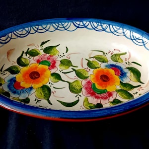 Könnte beinhalten: Eine ovale Keramik-Servierschale mit zwei seitlichen Griffen, die ein cremefarbenes Inneres mit handgemalten Blumenmotiven in Gelb, Rosa und Blau aufweist, umgeben von grünen Blättern und einem blauen, bogenförmigen Rand.