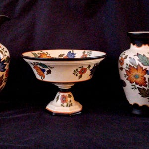 May include: A collection of ceramic vases and a pedestal bowl. The pieces are white with a floral design in orange, blue, and green. The pitcher has a handle and a black rim. The bowl is on a pedestal base.