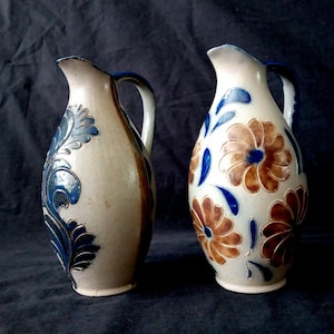 Op de afbeelding: Twee keramische kruiken met handvatten. Eén kruik heeft blauwe bloemmotieven, de andere blauwe bladeren. Beide hebben een crèmekleurige basis en bruine accenten. De kruiken worden tegen een donkere achtergrond getoond.