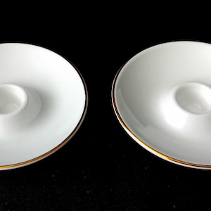Op de afbeelding: Twee witte porseleinen eierdopjes met een ronde voet en een centrale uitsparing voor een ei. Elk item is voorzien van een dunne, metallic gouden rand langs de buitenkant van het schoteltje, wat zorgt voor een strak en eenvoudig ontwerp.