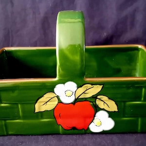 May include: Green ceramic basket with a handle, featuring a red apple and white flower design. The basket has a rectangular shape with a woven brick pattern and a gold trim around the edges. A decorative piece for the kitchen.