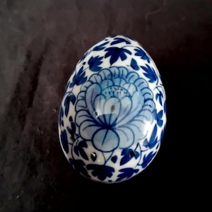 Op de afbeelding: Een decoratief keramisch ei met een blauw en wit bloemmotief. Het ei heeft een groot blauw bloemontwerp op de voorkant, omgeven door blauwe bladeren op een witte achtergrond. Het ei is ongeveer 7,6 cm hoog.