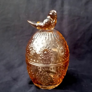 Op de afbeelding: Een amberkleurige glazen pot met een deksel voorzien van een handvat in de vorm van een vogel. De pot heeft een ovale vorm en is versierd met een verhoogd, reliëfpatroon van cirkelvormige bloemmotieven en kleine stippen over het gehele oppervlak.