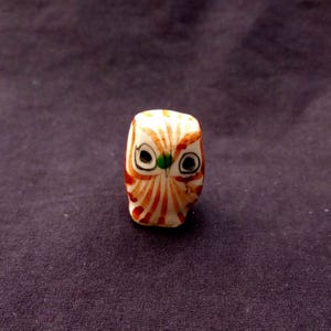 Op de afbeelding: Een kleine, handgeschilderde uilfiguur. De uil heeft een witte basis met oranje en rode details, waaronder strepen en gezichtskenmerken. De ogen zijn zwart met groene pupillen. Het beeldje staat op een donkere achtergrond.