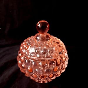 Könnte beinhalten: Eine rosa Bonbonniere aus Glas mit Deckel. Die Schale hat ein strukturiertes, blasenartiges Muster, das ihre Oberfläche bedeckt. Der Deckel ist mit einem kleinen, runden Knauf versehen. Die Schale steht vor einem dunklen Hintergrund.
