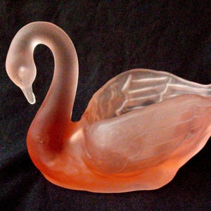 Op de afbeelding: Een zwaanbeeldje van matglas in roze en perziktinten. De zwaan heeft een gebogen nek en een gedetailleerd vleugelontwerp. Het beeldje staat tegen een donkere achtergrond.